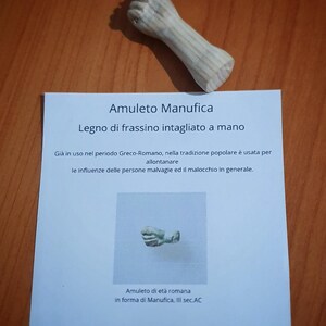 May include: A carved wooden amulet in the shape of a fist, with a detailed, textured surface. The amulet is accompanied by a card with Italian text, including the words "Amuleto Manufica" and "Legno di frassino intagliato a mano."