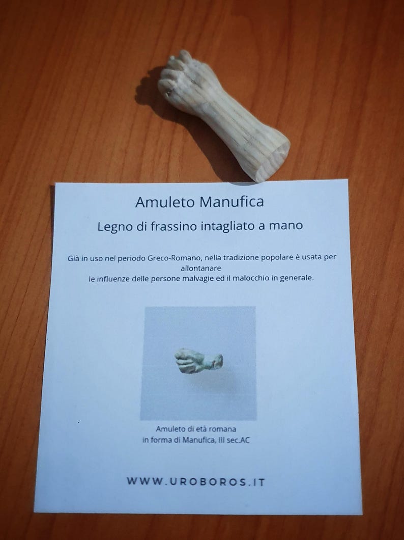 May include: A carved wooden amulet in the shape of a fist, with a detailed, textured surface. The amulet is accompanied by a card with Italian text, including the words "Amuleto Manufica" and "Legno di frassino intagliato a mano."