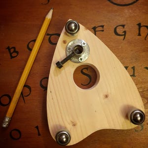 Può includere: Una planchette di legno con cuscinetti a sfera in metallo e un foro centrale, appoggiata su una superficie di legno scuro con lettere e numeri. Una matita gialla è accanto. La planchette ha un bullone e una rondella di metallo.