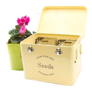 May include: A yellow metal seed storage box with a hinged lid and two clasps. The box has the text "Grow Your Own Seeds Storage Box" printed on the front. The box is open and contains several small packets of seeds.