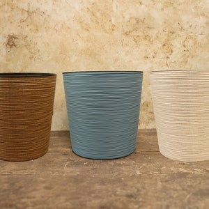May include: Three cylindrical planters with a textured, horizontal-line design. The planters are brown, blue-grey, and off-white, with black rims. They sit on a wooden surface against a mottled background.