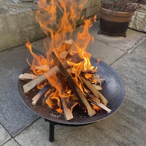 May include: A metal fire pit with a round bowl and three legs. The fire pit is filled with burning wood and flames.
