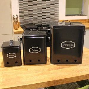 May include: Three black metal storage containers with labels for potatoes, onions, and garlic. The containers have holes in the bottom for ventilation.