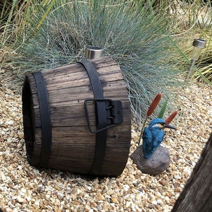 May include: A weathered wooden barrel with black metal bands and a handle. A blue and teal kingfisher statue sits on a rock. The barrel is surrounded by pebbles and greenery.
