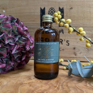Può includere: Una bottiglia di vetro marrone con tappo dorato, etichettata "HAND POURED IN SCOTLAND" e "LIME, BASIL & ORANGE SCENTED WITH ESSENTIAL OILS." La bottiglia è su una superficie di legno, con un fiore viola e bacche gialle sullo sfondo.