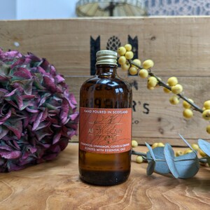 Puede incluir: Una botella de vidrio marrón con tapa plateada y una etiqueta naranja que dice "Hand Poured in Scotland" y "Med Oook Apothecary". La etiqueta enumera los aromas: "Madera de cedro, canela, clavo y naranja perfumados con aceites esenciales".