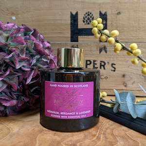 Puede incluir: Una botella de vidrio marrón oscuro con tapa dorada y etiqueta rosa. La etiqueta dice "Hand Poured in Scotland" y "Geranium, Bergamot & Lavender Scented with Essential Oils". La botella está junto a una flor morada y una caja de madera.