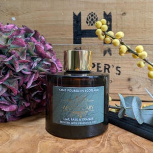 May include: A dark brown apothecary jar with a gold lid, labeled "Hand Poured in Scotland." The label reads "Lime, Basil & Orange Scented with Essential Oils." The jar is set against a wooden background with purple flowers and yellow berries.