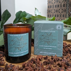 Puede incluir: Una vela de vidrio marrón con una etiqueta azul verdosa que dice "Melt Pool Apothecary Ylang Ylang & Patchouli Soy Wax Candle". La vela está sentada sobre una superficie de madera con vainas de anís estrellado esparcidas a su alrededor. La vela está al lado de una caja azul verdosa con la misma etiqueta y el texto "Hand Poured in Scotland 100% Natural Ingredients".