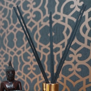 Könnte beinhalten: Schwarze Diffusorstäbchen in einer goldenen Glasflasche. Die Stäbchen sind V-förmig angeordnet. Die Flasche steht auf einem Tisch mit gemustertem Hintergrund.