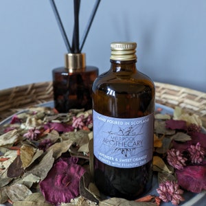 Puede incluir: Una botella de vidrio marrón con tapa dorada y una etiqueta que dice "MELT POOL APOTHECARY LAVENDER & SWEET ORANGE SCENTED WITH ESSENTIAL OILS". Un difusor de caña y botánicos secos rodean la botella. También es visible el texto "HAND POURED IN SCOTLAND".