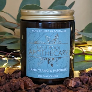 Op de afbeelding: Een bruine glazen kaars pot met een gouden deksel. Het etiket is blauw met gouden letters en luidt "Melt Pool Apothecary Ylang Ylang & Patchouli Soy Wax Candle".