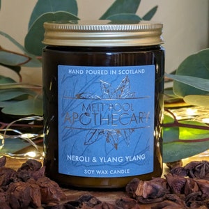 May include: A brown glass candle jar with a gold lid and a blue label. The label says "Melt Pool Apothecary" and "Neroli & Ylang Ylang Soy Wax Candle". The candle is sitting on a wooden surface with star anise.