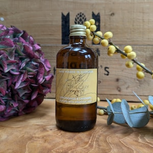 Puede incluir: Una botella de vidrio marrón con tapa dorada y etiqueta crema. La etiqueta dice "MELT POOL OTHECARY" y "LEMONGRASS & LAVENDER SCENTED WITH ESSENTIAL OILS." La botella está junto a flores moradas y bayas amarillas.