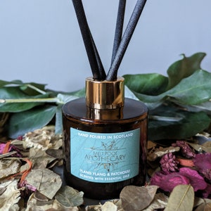 Op de afbeelding: Een bruine glazen diffuser met een gouden dop en zwarte rietjes. Het etiket vermeldt "HAND POURED IN SCOTLAND MELT POOL APOTHECARY YLANG YLANG & PATCHOULI SCENTED WITH ESSENTIAL OILS".