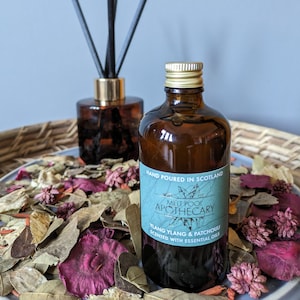 Op de afbeelding: Een bruine glazen fles met een gouden dop en een blauw etiket met de tekst "HAND POURED IN SCOTLAND MELT POOL APOTHECARY YLANG YLANG & PATCHOUU SCENTED WITH ESSENTIAL OILS".