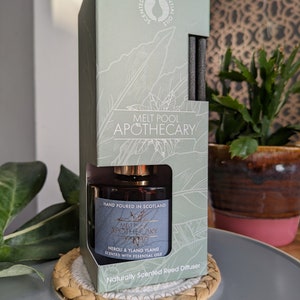 May include: A green and gold box with a reed diffuser bottle inside. The box has a white circle with the text "SCENTED WITH ESSENTIAL OILS" and the text "MELT POOL APOTHECARY". The bottle is labeled "MELT POOL APOTHECARY NEROI & YLANG YLANG SCENTED WITH ESSENTIAL OILS" and "HAND POURED IN SCOTLAND". The bottle is sitting on a woven round mat.