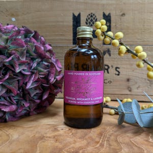 Op de afbeelding: Een bruine glazen fles met een gouden dop en een roze etiket met de tekst "Hand Poured in Scotland". Het etiket vermeldt ook "Verdant Apothecary" en vermeldt geranium, bergamot en lavendel. De fles staat naast een paarse bloem en een tak met gele bessen.