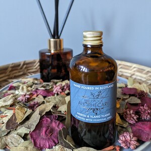 Può includere: Una bottiglia di vetro marrone con un tappo dorato e un'etichetta blu che recita "HAND POURED IN SCOTLAND MELT POOL APOTHECARY NEROI & YLANG YLANG SCENTED WITH ESSENTIAL OILS". La bottiglia è appoggiata su un letto di petali di fiori secchi.