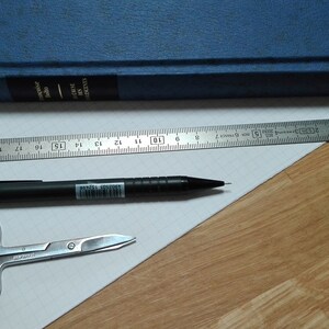 May include: A white sheet of paper with a black pencil, a pair of silver scissors, and a silver ruler. The ruler is 20 inches long and has markings for each inch. The paper is on a wooden surface.