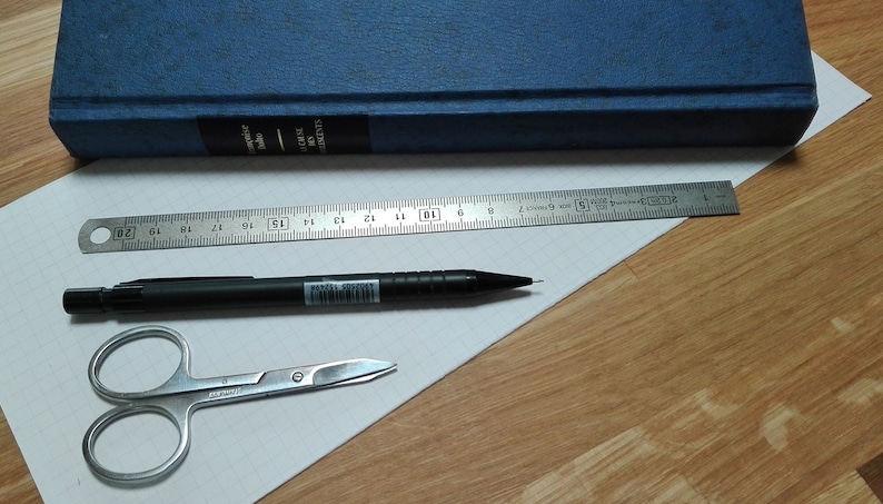 May include: A white sheet of paper with a black pencil, a pair of silver scissors, and a silver ruler. The ruler is 20 inches long and has markings for each inch. The paper is on a wooden surface.