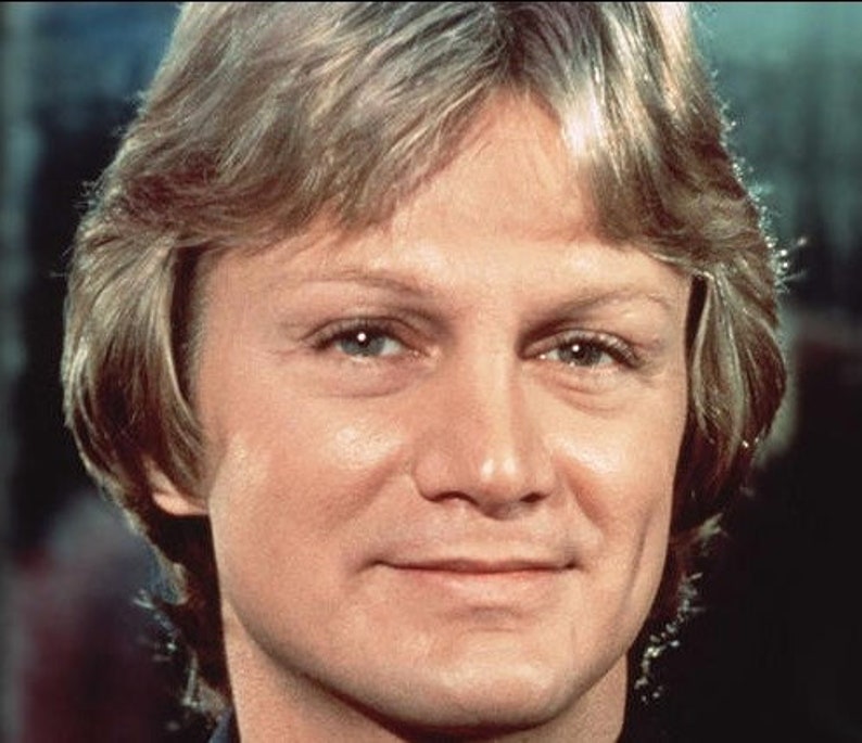 May include: A close-up portrait of a man with light brown hair, blue eyes, and a light complexion. He is smiling slightly and has a light brown beard.