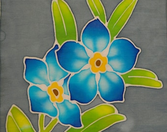 Puede incluir: Un panel de tela cuadrado con un diseño batik pintado a mano de dos flores nomeolvides azules con centros amarillos y hojas de color verde brillante sobre un fondo gris apagado. Una etiqueta blanca en la esquina dice FORGET ME NOT 1 www.turtlehand.com.