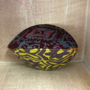 May include: A round, colorful pillow with a geometric pattern in shades of red, blue, and green. The pillow is made of fabric and has a sewn seam.
