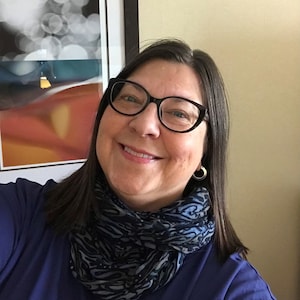 May include: A woman with brown hair wearing black cat-eye glasses and a blue patterned scarf. She is smiling and looking at the camera.