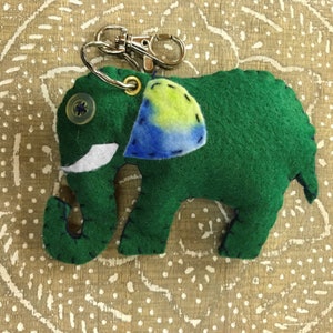 Può includere: Portachiavi a forma di elefante in feltro verde con una toppa in feltro blu e giallo sul lato. L'elefante ha una zanna in feltro bianco e un bottone nero per l'occhio.