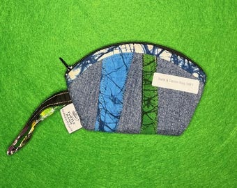 Puede incluir: Un pequeño bolso Batik & Denim Bag SM1 hecho a mano, con cierre de cremallera y una correa de muñeca estampada. El bolso presenta paneles de tela azul, verde y denim. El fondo es de color verde liso.