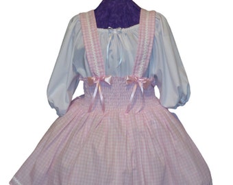 Può includere: Un vestito a quadri vichy rosa e bianco con bretelle bianche e fiocchi rosa. Il vestito ha un bordo di pizzo bianco e una camicetta bianca con un fiocco.