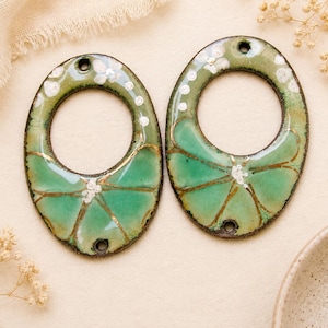 Peut inclure: Deux pendentifs ovales en émail faits à la main. Les pendentifs sont verts avec des accents dorés et des détails floraux blancs. Chaque pendentif a un trou en haut et en bas pour l'enfilage. Le fond est beige neutre.