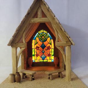 May include: A miniature wooden chapel with a stained-glass window. The chapel features a peaked roof, two benches, and two small vases. The stained-glass window is illuminated, showcasing a colorful floral design with red, blue, and yellow hues.
