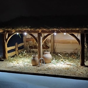 May include: A miniature wooden nativity scene with a thatched roof, a hay-filled stable, and two clay pots. The scene is lit with small white lights.