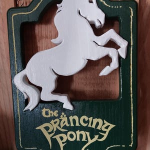 Könnte beinhalten: Holzschild mit einem weißen Pferd in tänzerischer Pose, vor einem dunkelgrünen Hintergrund. Das Schild trägt die Aufschrift "The Prancing Pony" in goldfarbenen Buchstaben mit einem goldenen Rand. Enthält zwei Metallhaken zum Aufhängen.