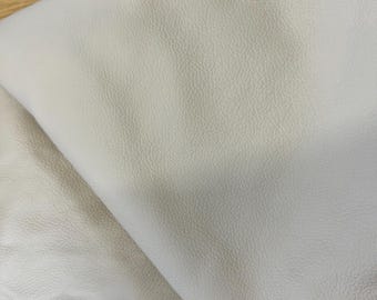 May include: A piece of off-white leather with a visible grain texture. The material is folded over itself, showing the smooth, matte surface on the top side and a lighter, suede-like texture on the underside of the leather material.