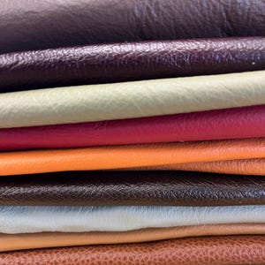 Könnte beinhalten: Ein Stapel gefalteter Lederstücke in verschiedenen Farben, darunter Dunkelbraun, Dunkelviolett, Hellgrün, leuchtendes Pink, kräftiges Orange, Schwarz, Hellblau, Beige und strukturiertes Hellbraun. Jedes Stück zeigt eine eigene Narbung und matte Oberfläche.