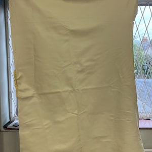 Puede incluir: Una gran piel de cuero amarillo pálido cuelga de pinzas, llenando el marco. La piel tiene una superficie lisa, ligeramente texturizada y está drapeada en suaves pliegues. Los bordes son irregulares, lo que sugiere un corte natural. Se ve una ventana al fondo.