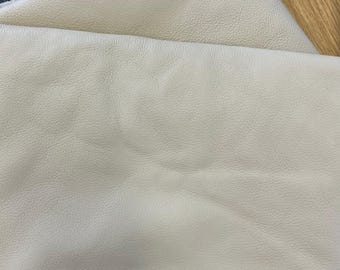 May include: A folded piece of off-white leather featuring a natural, pebbled grain texture. The material displays soft, organic folds and creases across its surface, highlighting the flexible and pliable nature of the light-colored hide.