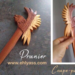 May include: A wooden bird-shaped paper knife with a reddish-brown body and light brown wings. The knife is carved with intricate details and has a sharp point. The text "Prunier" and "www.ehlyass.com" is printed below the knife. The text "Coupe-papier" is printed below the knife in the right corner of the image.
