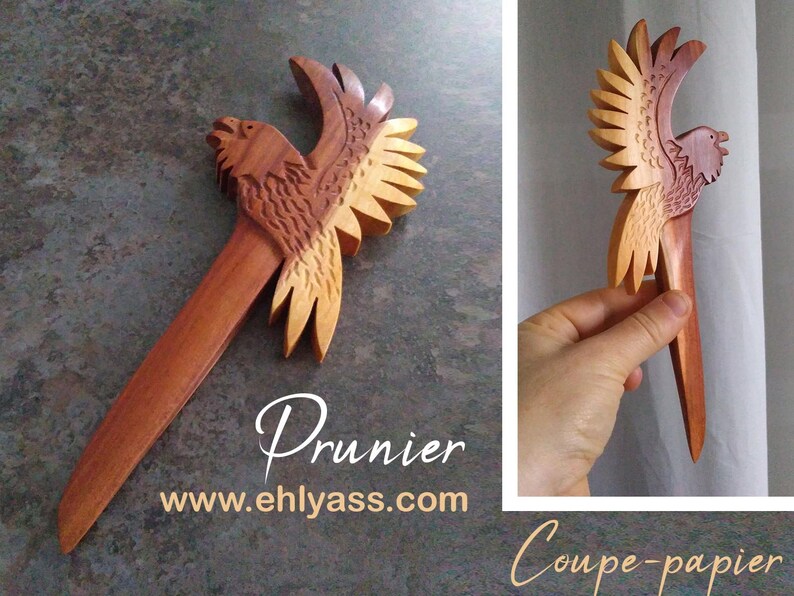 May include: A wooden bird-shaped paper knife with a reddish-brown body and light brown wings. The knife is carved with intricate details and has a sharp point. The text "Prunier" and "www.ehlyass.com" is printed below the knife. The text "Coupe-papier" is printed below the knife in the right corner of the image.