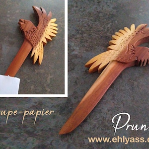 May include: A wooden letter opener shaped like a bird with its wings spread. The bird is carved in a light brown wood with darker brown accents. The letter opener is about 12 inches long. The text "Coupe-papier" and "Prunier" are visible in the image.