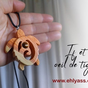Puede incluir: Un colgante de madera con forma de tortuga con una piedra de ojo de tigre en el centro. El colgante está en un cordón negro y se sostiene en una mano. El texto "T et oeil de tigre" y "www.ehlyass.com" son visibles en el fondo.