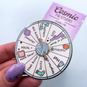 May include: A round enamel pin with a white background and a spinning wheel design. The wheel features knitting-related icons and text, including "Sweater," "Mittens," and "Socks." The pin has a silver border and a knitting needle and yarn in the center.