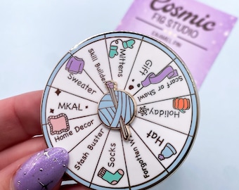 Könnte beinhalten: Runder Emaille-Pin mit einem Design eines Spinnrads. Er zeigt strickbezogene Symbole und Text wie "Sweater", "Mittens" und "Socks". Der Pin hat einen silbernen Rand und eine Stricknadel und Wolle in der Mitte.