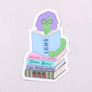 May include: A sticker featuring a green bookworm with purple hair and glasses reading a book labeled "SMUT." The worm is perched atop a stack of books with titles like "Enemies to Lovers" and "Slow Burn."