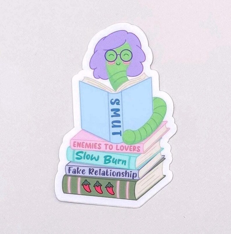 May include: A sticker featuring a green bookworm with purple hair and glasses reading a book labeled "SMUT." The worm is perched atop a stack of books with titles like "Enemies to Lovers" and "Slow Burn."