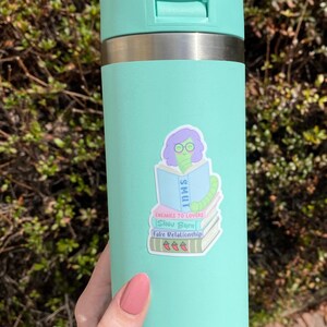 May include: A mint green Yeti water bottle with a matching handle and lid. A sticker on the bottle features a cartoon worm wearing glasses and reading a book labeled "SMUT". The sticker also includes the phrases "Enemies to Lovers", "Slow Burn", and "Fake Relationship".