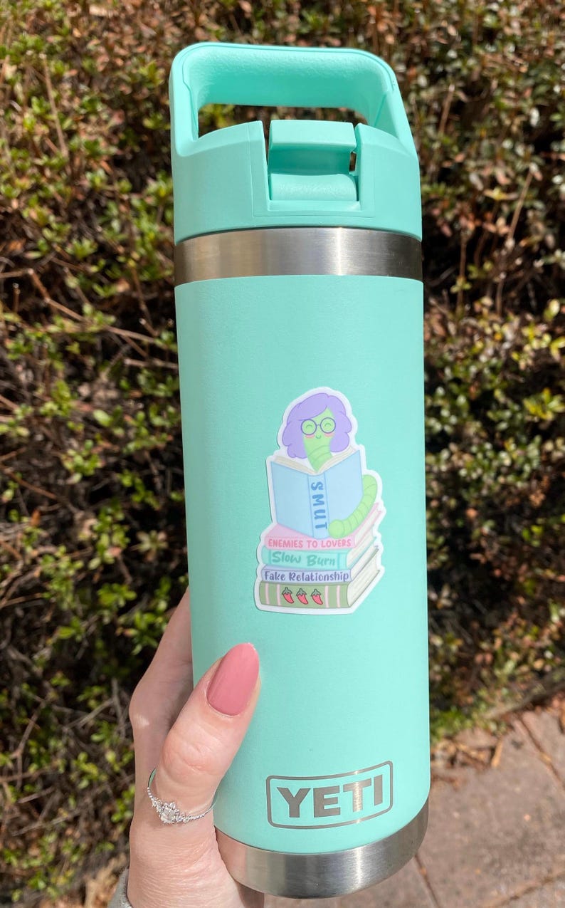 May include: A mint green Yeti water bottle with a matching handle and lid. A sticker on the bottle features a cartoon worm wearing glasses and reading a book labeled "SMUT". The sticker also includes the phrases "Enemies to Lovers", "Slow Burn", and "Fake Relationship".